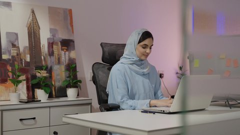 مهنة ووظيفة نسائية،  ادارة الأعمال وانجاز المهام المطلوبة، ايماءات التركيز والاندماج، ادارة وانجاز المهام باستخدام جهاز تقني حديث ومتطور، إمرأة عربية خليجية اماراتية محجبة ترتدي عباءة تجلس على المكتب تستخدم الحاسوب