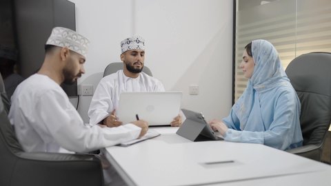 إدارة و تقسيم المهام، فريق عمل عربي خليجي عماني يرتدون الزي التقليدي يقومون بإجراء اجتماع عمل داخل غرفة الاجتماعات في مقر الشركة، كتابة و تدوين الملاحظات, دمج الأجهزة التقنية الحديثة بالأعمال المكتبية، دراسة أوراق البيانات الإحصائية, تطوير المشاريع و الاستثمارات