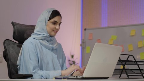 ادارة الأعمال وانجاز المهام المطلوبة، ايماءات التركيز والاندماج، ادارة وانجاز المهام باستخدام جهاز تقني حديث ومتطور، مهنة ووظيفة نسائية، تصوير مقرب من الجانب لإمرأة عربية خليجية اماراتية محجبة ترتدي عباءة تجلس على المكتب تستخدم الحاسوب