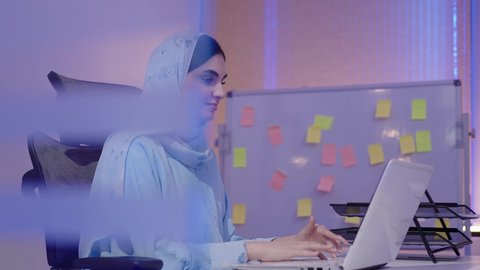 ايماءات التركيز والاندماج، ادارة وانجاز المهام باستخدام جهاز تقني حديث ومتطور، ادارة الأعمال وانجاز المهام المطلوبة، مهنة ووظيفة نسائية، تصوير مقرب من الجانب لإمرأة عربية خليجية اماراتية محجبة ترتدي عباءة تجلس على المكتب تستخدم الحاسوب