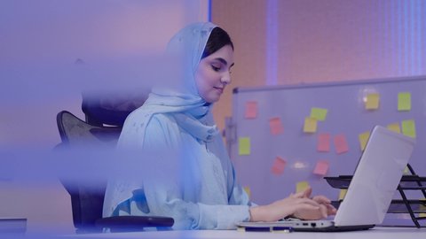 Managing and accomplishing tasks using a modern and advanced technical device, gestures of focus and engagement, business management and completing required tasks, a profession and job for women, a close-up side shot of an Arab Gulf Emirati woman wearing a hijab and an abaya sitting at a desk using a computer.