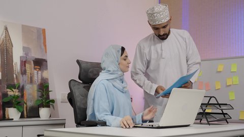 كتابة و تدوين الملاحظات، دمج التقنيات الحديثة المتطورة في الحياة العملية، مهن ووظائف مكتبية، امراة عربيه خليجيه عمانية ترتدي الحجاب و العباءة خلف مكتبها ويقوم بالعمل على جهاز الاب توب المحمول بمساعده زميلها لدراسه وتحليل المشاريع الجديده ، مناقشه اداء وتقارير العمل