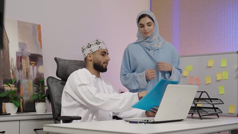 مهن ووظائف مكتبية، رجل عربي خليجي عمانى يرتدي الدشداشة و الكمة يجلس خلف مكتبه ويقوم بالعمل على جهاز الاب توب المحمول بمساعده زميلته لدراسه وتحليل المشاريع الجديده ، مناقشه اداء وتقارير العمل، دمج التقنيات الحديثة المتطورة في الحياة العملية