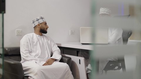 التفاعل الجماعي والوحدة لتحقيق أهداف الشركة وحل المشكلات، الإشارة إلى شاشة الحاسوب، مفهوم الإدارة المتعاونة، رجلان عربيان خليجيان عمانيان يرتديان الدشداشة و الكمة يناقشان أمور العمل داخل المكتب، استخدام التقنيه الحديثة