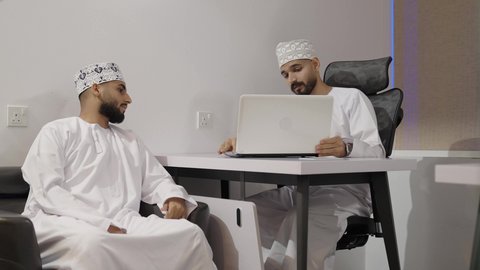 الإشارة إلى شاشة الحاسوب، التفاعل الجماعي والوحدة لتحقيق أهداف الشركة وحل المشكلات، مفهوم الإدارة المتعاونة، رجلان عربيان خليجيان عمانيان يرتديان الدشداشة و الكمة يناقشان أمور العمل داخل المكتب، استخدام التقنيه الحديثة