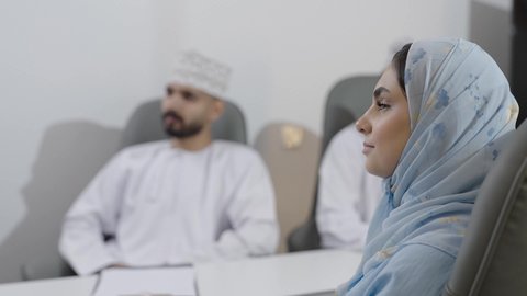 تعابير الاندماج و التركيز، توضيح و شرح المهام المطلوبة، تبادل الخبرات والمعلومات الناجحة، تصوير مقرب من الجانب لامراة عربية خليجيه عمانية ترتدي العباءة و الحجاب تجلس في غرفة الاجتماع داخل مقر الشركة برفقه زملائها