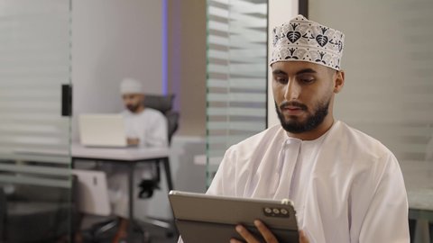 استعمال التكنولوجيا في العمل عن بعد، متابعة مواقع التواصل الاجتماعي عن طريق الانترنت، استخدام الأجهزة الحديثة في انجاز الأعمال، تصوير مقرب لرجل عربي خليجي عماني يرتدي الدشداشة و الكمة يحمل بيده جهاز لوحي في المكتب، ايماءات السعادة و السرور
