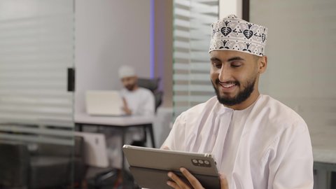 متابعة مواقع التواصل الاجتماعي عن طريق الانترنت، استخدام الأجهزة الحديثة في انجاز الأعمال، استعمال التكنولوجيا في العمل عن بعد،تصوير مقرب لرجل عربي خليجي عماني يرتدي الدشداشة و الكمة يحمل بيده جهاز لوحي في المكتب، ايماءات السعادة و السرور