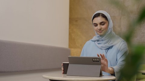 Browsing social media sites, enjoying drinks, using modern technology devices for work, the concept of remote project management, an Arab Gulf Omani woman in a hijab wearing an abaya completing work tasks via a tablet, secure online shopping.