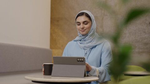 Integrating technology into daily and professional life, enjoying beverages, using modern technological devices for work, the concept of remote project management, an Arab Gulf Omani woman in a hijab wearing an abaya completing work tasks via a tablet, secure online shopping.
