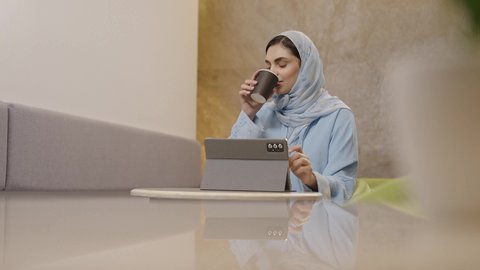 Enjoying beverages, using modern technology devices for work, the concept of remote project management, an Arab Gulf Omani woman in a hijab wearing an abaya completing work tasks via a tablet, secure online shopping, integrating technology into daily and professional life.