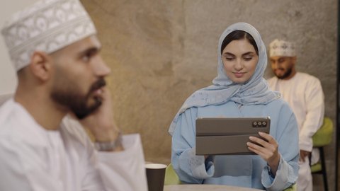 The use of modern technological devices for work, the concept of remote project management, an Arab Gulf Omani woman in a hijab wearing an abaya completing work tasks via a tablet, secure online shopping, a close-up shot of an Arab Gulf Omani man wearing a dishdasha and kumma making a phone call via a mobile phone.