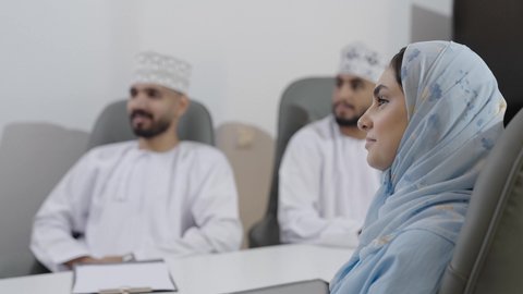 Clarification and explanation of the required tasks, exchange of successful experiences and information, gestures of happiness and joy, a close-up side shot of an Omani Gulf Arab woman wearing an abaya and hijab sitting in a meeting room inside the company headquarters with her colleagues.