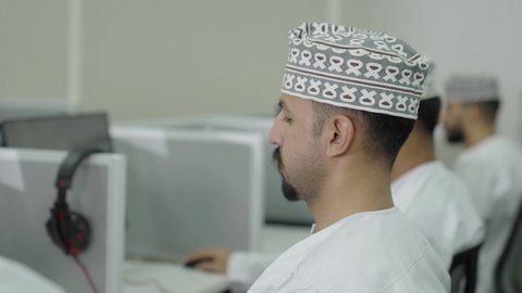 الدراسة المستمرة والعمل الجاد، التعليم في سلطنه عمان, تصوير مقرب من الجانب لطالب عربي خليجي عماني يرتدي الدشداشة و الكمة يجلس فى المحاضرة يقوم بوضع سماعات الراس على اذنه, ايماءات الاندماج و التركيز