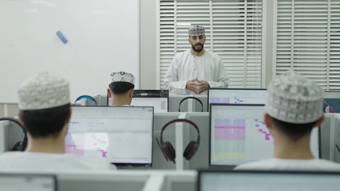 مفهوم التعليم بالمناهج المقررة، تصوير من الخلف لمجموعه من الطلاب يجلسون امام الحاسب المحمول يستمعون الى المحاضرة بايماءات الاندماج و التركيز، التعليم في سلطنه عمان، استخدام التقنية الحديثة، معلم عربي خليجي عماني يرتدي الدشداشة و الكمة يقوم بشرح الدرس للطلاب