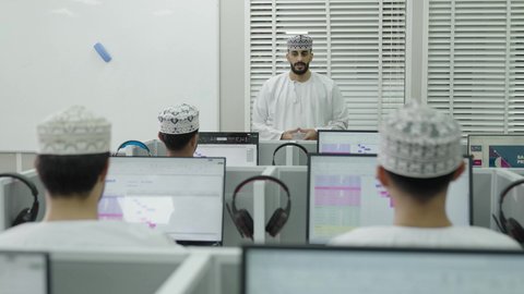 The concept of education in the Sultanate of Oman, the use of modern technology, an Arab Gulf Omani teacher wearing a dishdasha and kumma explaining the lesson to the students, a shot from behind of a group of students sitting in front of a laptop listening to the lecture with gestures of engagement and focus.