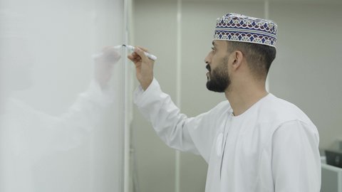 ، كتابة وتدوين الملاحظات على بورد ابيض، اقامة دورات تدريبية في عمان، تصويرمقرب من الجانب لمعلم عربي خليجي عاماني يرتدي الدشداشة و الكمة يقوم بالكتابة على السبورة، مهنة المعلم في الجامعات التعليمية