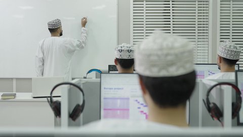 الكتابة على بورد أبيض، اقامة دورات تدريبية في عمان، تصوير من الخلف لمعلم عربي خليجي عاماني يرتدي الدشداشة و الكمة يقوم بالكتابة على السبورة، مهنة المعلم في الجامعات التعليمية، مجموعه من الطلاب يقومون بالاستماع للمحاضرة