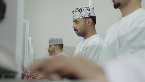 متابعة منصات التعلم الالكتروني بإستعمال اللابتوب، دمج التكنولوجيا بالتعليم، تصوير مقرب لطلاب عربيون خليجيون عمانيون يرتدون الدشداشة و الكمة يقومون بحل الواجبات اونلاين، النقر على لوحه المفتايح ، ايماءات الاندماج و التركيز
