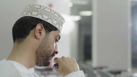The pursuit of excellence, perseverance, and a commitment to success are gestures that indicate focus and contemplation of the answer. A close-up side shot of an Omani Gulf Arab student wearing a dishdasha and kumma solving exam questions.
