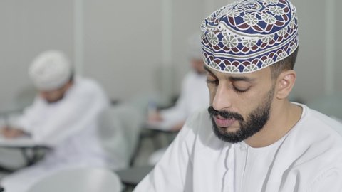 Expressions of engagement and focus, a close-up shot of an Omani Gulf Arab student wearing a dishdasha and kumma looking at something, a group of students solving an exam.