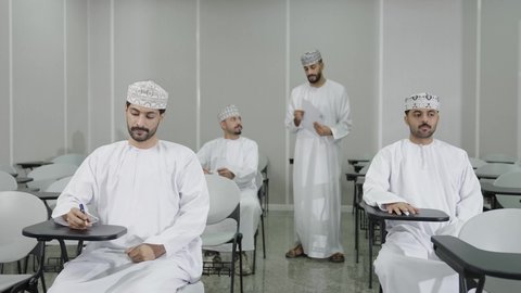 التعليم في سلطنه عمان، الدراسة و الاجتهاد من اجل التفوق, مجموعة من التلاميذ العمانيين يقومون بالاستعداد لحل أسئلة الامتحان, اهداف التربية و التعليم, معلم عربي خليجي عماني يرتدي الدشداشة و الكمه يقوم بتوزيع ورق الامتحان على الطلاب