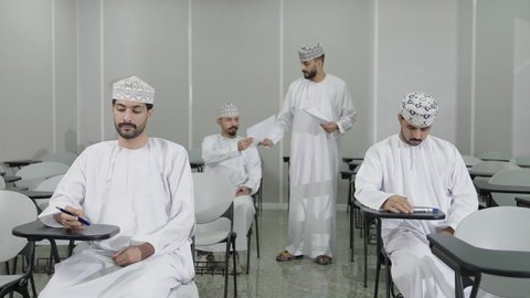 الدراسة و الاجتهاد من اجل التفوق, مجموعة من التلاميذ العمانيين يقومون بالاستعداد لحل أسئلة الامتحان, اهداف التربية و التعليم, التعليم في سلطنه عمان ,معلم عربي خليجي عماني يرتدي الدشداشة و الكمه يقوم بتوزيع ورق الامتحان على الطلاب