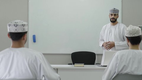 كليات وجامعات متطوره، أستاذ جامعي عربي خليجي عماني يرتدي الدشداشة و الكمة يشرح لطلابه في المحاضره, رفع اليدين للتفاعل والمشاركه, الإستماع الى المحاضره, جامعات أكاديميه في سلطنه عمان