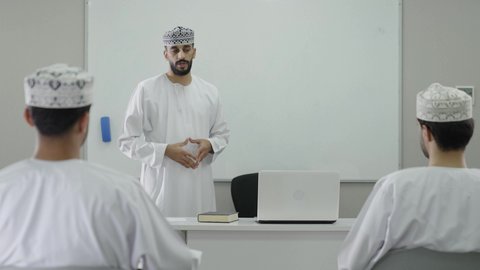 أستاذ جامعي عربي خليجي عماني يرتدي الدشداشة و الكمة يشرح لطلابه في المحاضره, رفع اليدين للتفاعل والمشاركه, الإستماع الى المحاضره, جامعات أكاديميه في سلطنه عمان , كليات وجامعات متطوره