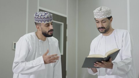 مناقشة المواد الدراسيه، فصل دراسي جديد، العمل الجماعي والتعاون بين الزملاء، تأدية الواجبات الجامعية، طالب عربي خليجي عماني يرتدي الدشداشة و الكمة يمسك بيديه كتاب يقوم بمشاركة المعلومات مع زميله، التعليم الجامعي في سلطنه عمان