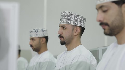 التعليم العالي في سلطنة عمان، تصوير مقرب  من الجانب لطلاب عربيون خليجيون عمانيون  يرتدون الدشداشة و الكمة يجلسون فى قاعة المحاضرات الجامعية، ايماءات الاندماج و التركيز