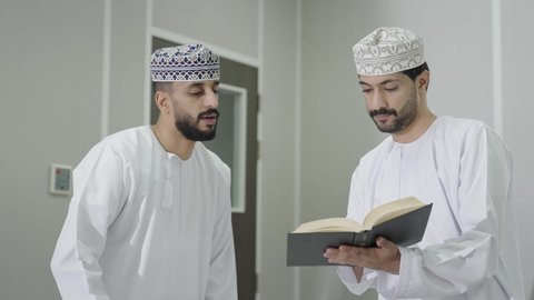 فصل دراسي جديد، العمل الجماعي والتعاون بين الزملاء، مناقشة المواد الدراسيه، تأدية الواجبات الجامعية، طالب عربي خليجي عماني يرتدي الدشداشة و الكمة يمسك بيديه كتاب يقوم بمشاركة المعلومات مع زميله، التعليم الجامعي في سلطنه عمان