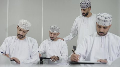 مراقبة الطلاب أثناء حل الامتحان، الدراسة والاجتهاد من أجل التفوق، معلم عربي خليجي عماني يرتدي الدشداشة و الكمه يقوم بشرح شيء ما لاحد التلاميذ، النجاح والانجاز في المرحلة الجامعية، ايماءات الاندماج و التركيز
