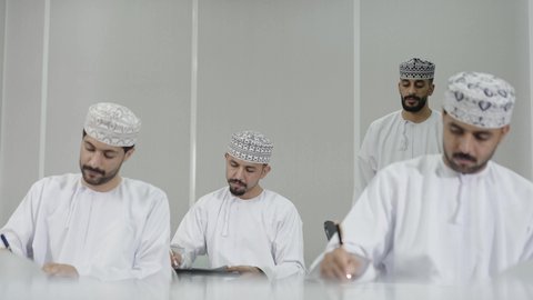 الدراسة والاجتهاد من أجل التفوق، مراقبة الطلاب أثناء حل الامتحان، معلم عربي خليجي عماني يرتدي الدشداشة و الكمه يقوم بشرح شيء ما لاحد التلاميذ، النجاح والانجاز في المرحلة الجامعية، ايماءات الاندماج و التركيز