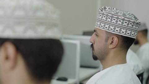 Studying and striving for excellence, education in the Sultanate of Oman, a close-up side shot of an Arab Gulf Omani student wearing a dishdasha and kumma sitting in a lecture, putting headphones on his ears, gestures of engagement and focus.
