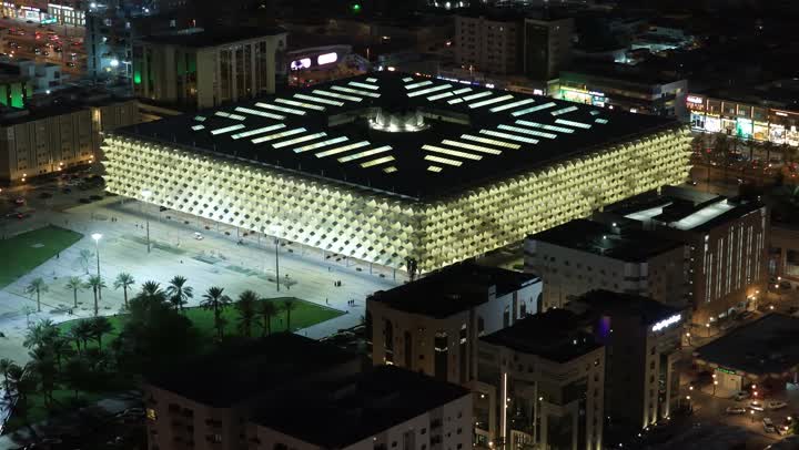 معالم الرياض الثقافية المشهورة في الليل، تصوير درون جوي للفن الهندسي المعماري الخارجي لمكتبة الملك فهد المضاءة  في مدينة الرياض، الأشجار والنباتات الخضراء على الطرق والشوارع العامة، المكتبات الوطنية في المملكة العربية السعودية