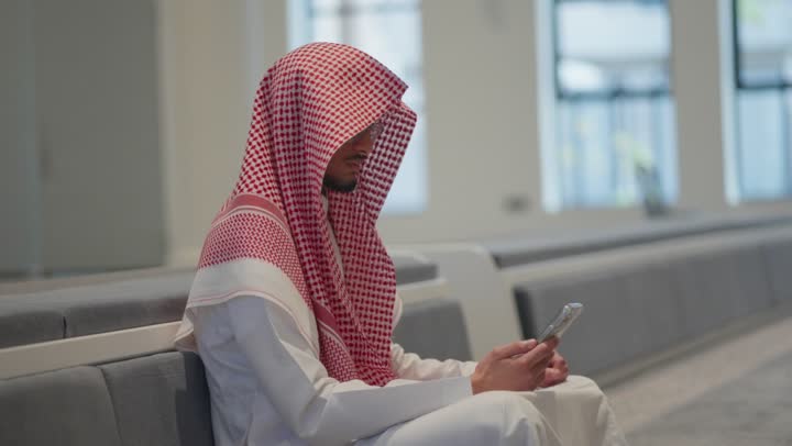 Reading the Holy Quran from a mobile phone, using a modern and advanced technical device, a Saudi Arabian Gulf man wearing traditional thobe and shemagh prostrating inside the mosque, worship and drawing closer to God, worship in the houses of God
