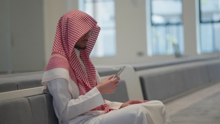 قراءة القران الكريم من الهاتف المحمول، استخدام جهاز تقني حديث ومتطور،  رجل عربي خليجي سعودي يرتدي الثوب التقليدي والشماغ يسجد داخل المسجد، العبادة و التقرب الى الله، التعبد في بيوت الله