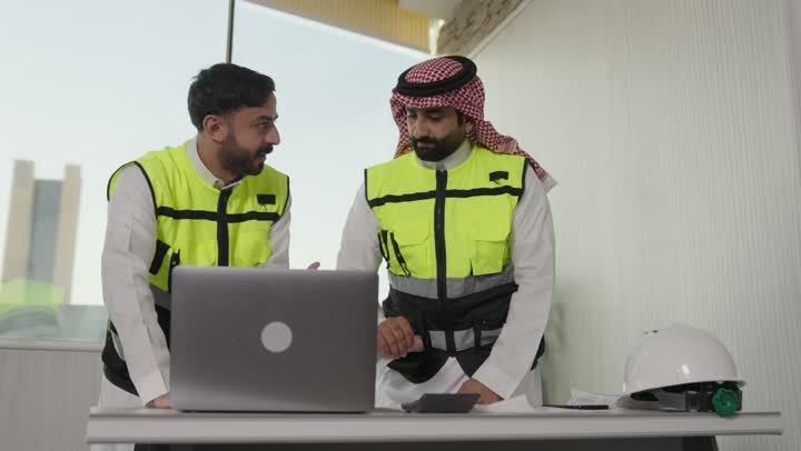 Integrating work with technology, reviewing new architectural designs on a laptop, an Arab Saudi Gulf architect wearing work attire uses a laptop alongside a colleague, overseeing the electronic application of architectural projects and plans in construction.
