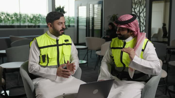 دمج العمل مع التقنية و التكنولوجيا، الاطلاع على تصاميم هندسية جديدة على جهاز الحاسوب المحمول، مهندس معماري عربي سعودي خليجي يرتدي اللباس الخاص بالعمل يستخدم اللابتوب برفقة زميله بالعمل ، الاشراف الالكتروني على تطبيق المشاريع و الخطط الهندسيه المعمارية في البناء