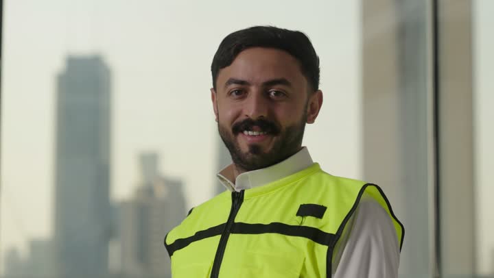 النظر الى الكاميرا والوقوف بإستقامة، تعابير السرور والسعادة والثقة بالنفس، العمل في القطاع الهندسي، مفهوم الهندسة والانشاءات المعمارية، بورتريه لمهندس عربي خليجي سعودي مبتسم يرتدي الزى التقليدي والشماغ وسترة الحماية