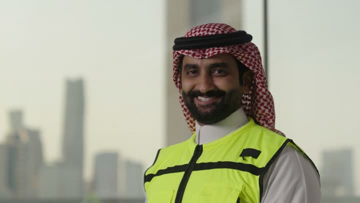 النظر الى الكاميرا والوقوف بإستقامة، تعابير السرور والسعادة والثقة بالنفس، العمل في القطاع الهندسي، مفهوم الهندسة والانشاءات المعمارية، بورتريه لمهندس عربي خليجي سعودي مبتسم يرتدي الزى التقليدي والشماغ وسترة الحماية