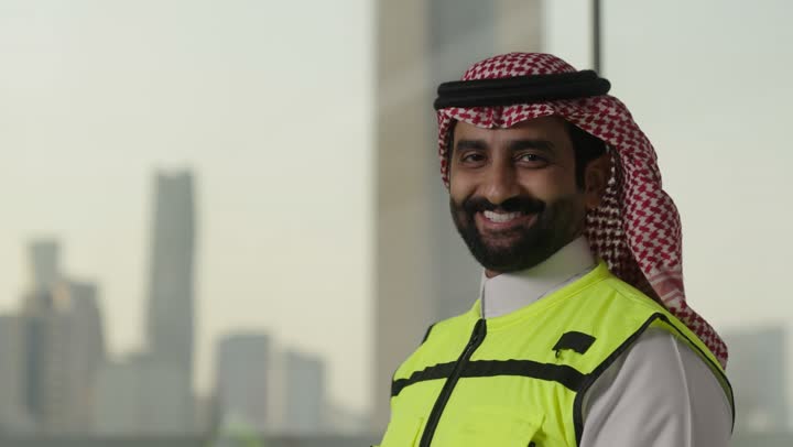 تعابير السرور والسعادة والثقة بالنفس، النظر جانباً والوقوف بإستقامة، العمل في القطاع الهندسي، مفهوم الهندسة والانشاءات المعمارية، بورتريه لمهندس عربي خليجي سعودي مبتسم يرتدي الزى التقليدي والشماغ وسترة الحماية