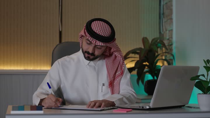 Gestures of integration and focus, working on a laptop inside the office, an administrative office profession and job, managing business affairs through a modern and advanced technical device, a Saudi Gulf Arab employee wearing traditional thobe and shemagh using a laptop and taking notes, companies and engineering offices in Saudi Arabia.