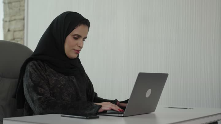 A profession and administrative office job, managing business affairs through a modern and advanced technical device, gestures of integration and focus, working on a laptop inside the office, an Arab Gulf Saudi female employee wearing an abaya and hijab using a laptop and taking notes, companies and engineering offices in Saudi Arabia.