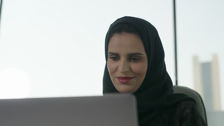 A profession and administrative office job, managing business affairs through a modern and advanced technical device, gestures of integration and focus, working on a laptop inside the office, an Arab Gulf Saudi female employee wearing an abaya and hijab using a laptop and taking notes, companies and engineering offices in Saudi Arabia.