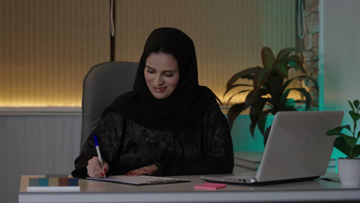 A profession and administrative office job, managing business affairs through a modern and advanced technical device, gestures of integration and focus, working on a laptop inside the office, an Arab Gulf Saudi female employee wearing an abaya and hijab using a laptop and taking notes, companies and engineering offices in Saudi Arabia.