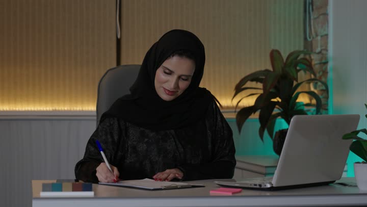 A profession and administrative office job, managing business affairs through a modern and advanced technical device, gestures of integration and focus, working on a laptop inside the office, an Arab Gulf Saudi female employee wearing an abaya and hijab using a laptop and taking notes, companies and engineering offices in Saudi Arabia.