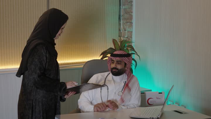 The concept of medicine and healthcare, managing business affairs through a modern and advanced technological device, working in the healthcare sector, working on a laptop, an Arab Gulf Saudi doctor wearing a white coat and stethoscope using a laptop and taking notes, the medical clinic in Saudi Arabia.