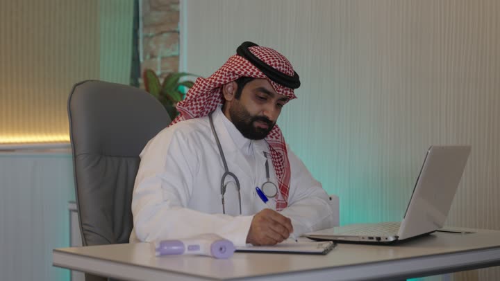 العمل فى القطاع الصحي ، مفهوم الطب والرعاية الصحية، إدارة شؤون الأعمال عبر جهاز تقني حديث ومتطور، ، العمل على الحاسوب المحمول  ،طبيب عربي خليجي سعودي يرتدي المعطف الابيض والسماعه  يقوم باستخدام الحاسوب المحمول وتدوين الملاحظاات ، العيادة الطبية في السعودية